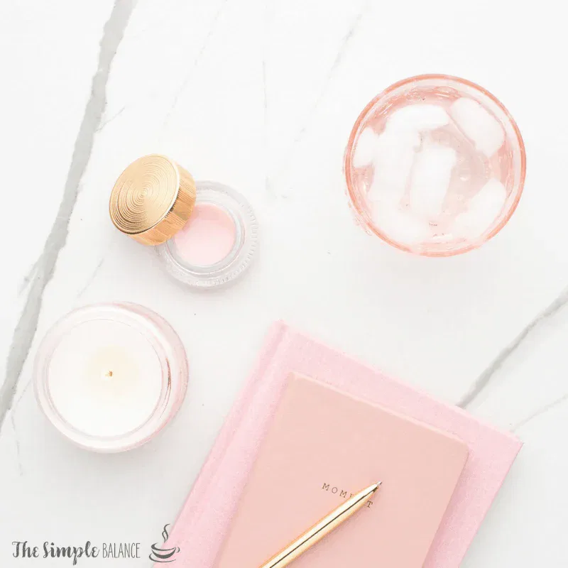 Calming self-care setup with pink candle, MOM notebook, golden pen, and refreshing drink on marble.